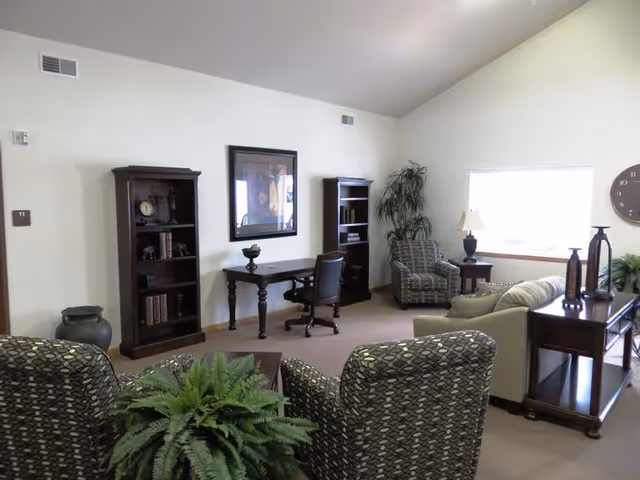 A well-lit living room area with two patterned armchairs, a beige sofa, a wooden desk with a chair, two tall bookshelves, a side table with a lamp, a large wall clock, and several decorative plants. The walls are light-colored and there is a window letting in natural light.