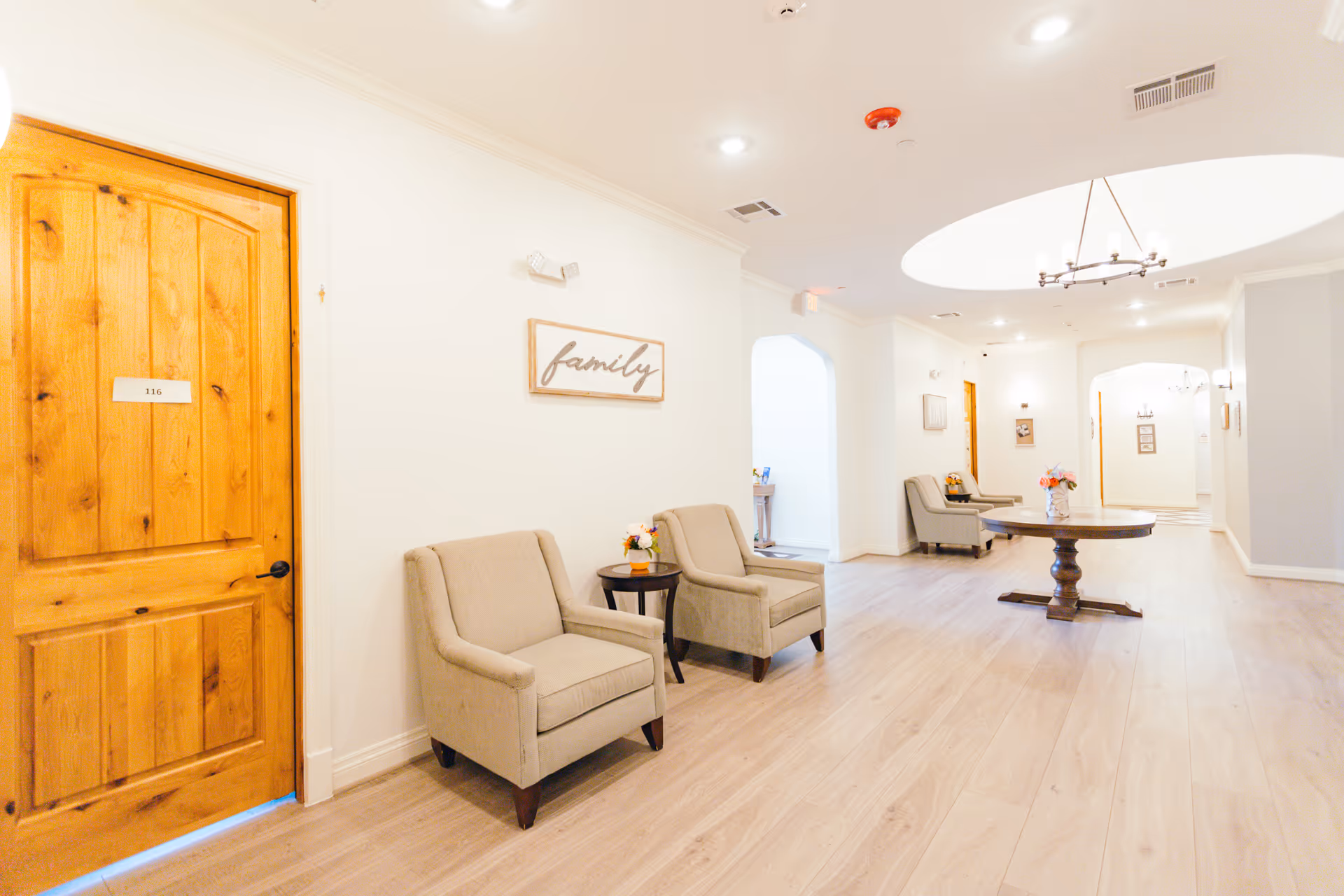 Bright, airy interior hallway/lounge with beige armchairs, a central round wooden table under a skylight, and a wooden door labeled 116.