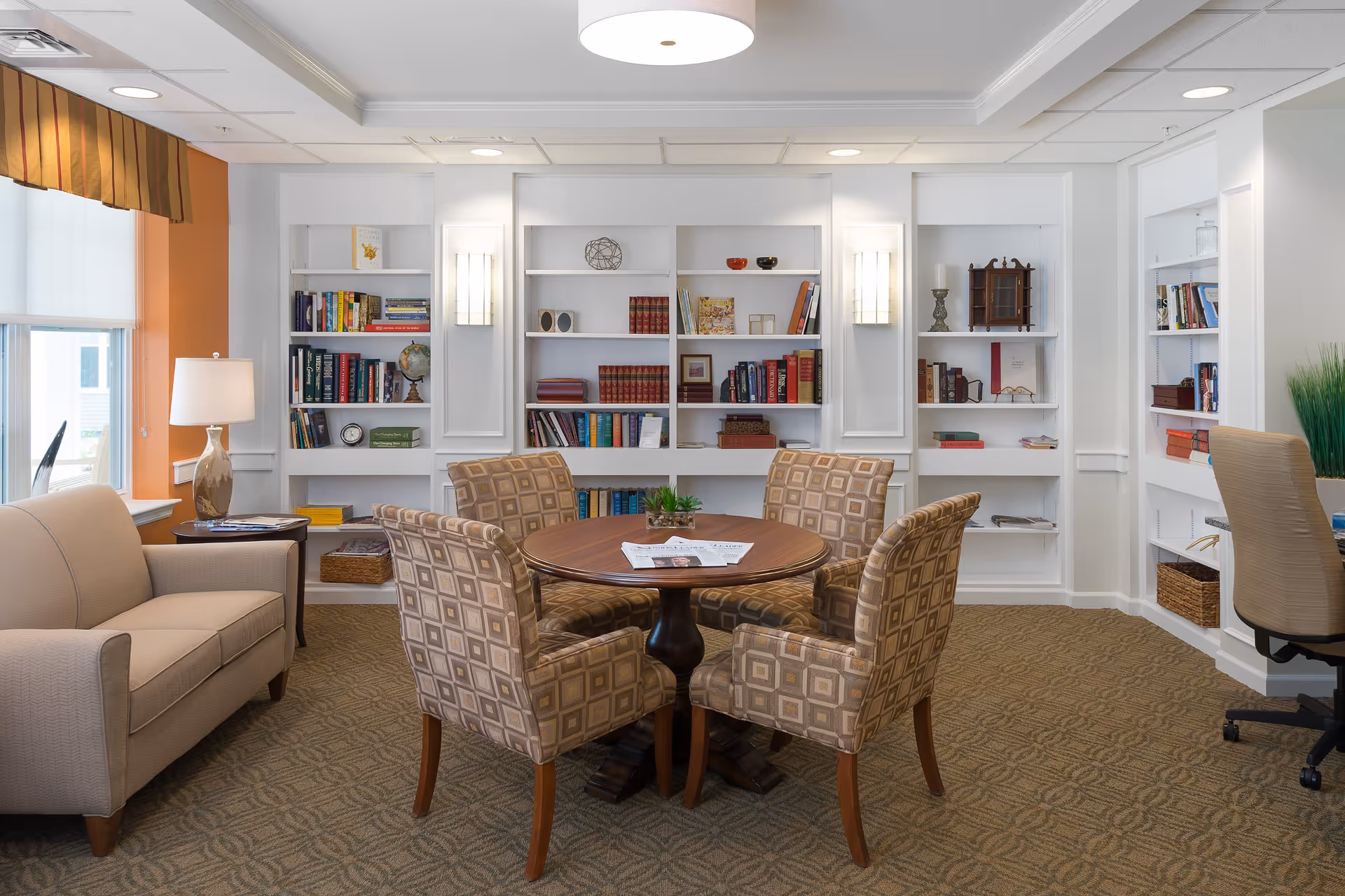 Bright common room with a round wooden table surrounded by four patterned chairs in front of built-in bookshelves, with a sofa and lamp by the window.