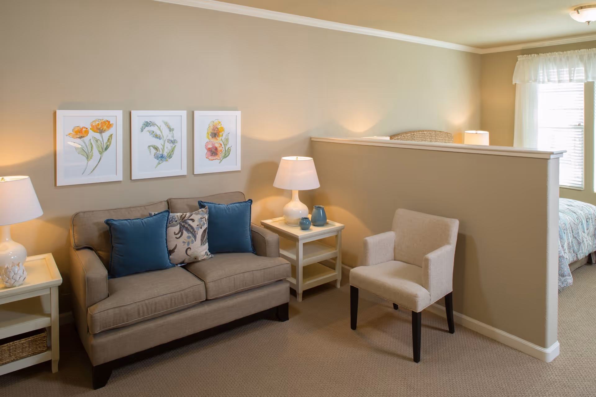 A cozy living area with a beige sofa adorned with blue and patterned cushions, a beige armchair, two white side tables each with a white lamp, and three framed floral paintings on the wall. A half wall separates the living area from a bedroom with a bed covered in a patterned bedspread and a window with sheer white curtains.