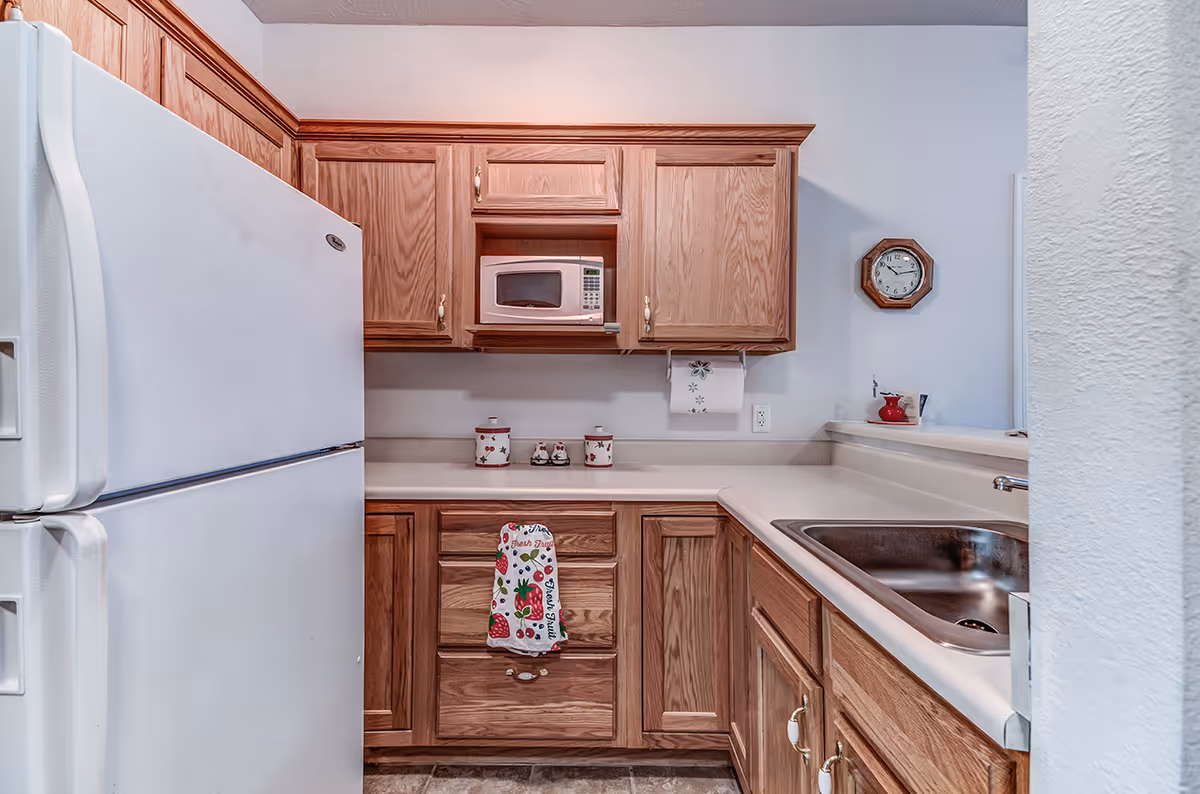 Small kitchen with oak cabinets, a white refrigerator, microwave, stainless sink, and countertop with decorative items.