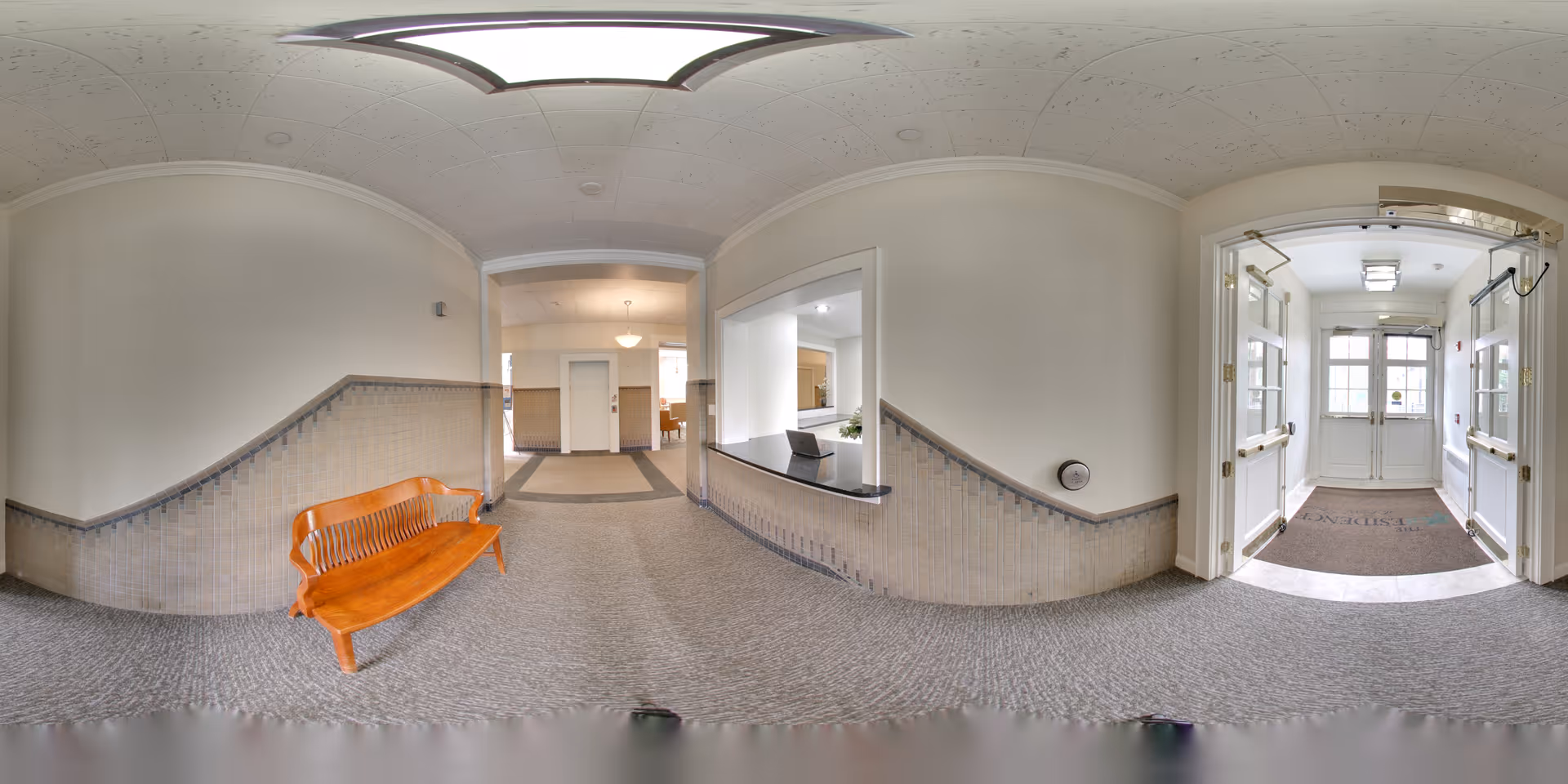 Interior view of a senior living facility hallway with beige walls and carpeted floor. A wooden bench is placed against the left wall. There is a reception window on the right side and double doors leading outside at the far end. The area is well-lit with ceiling lights and natural light from the doors.