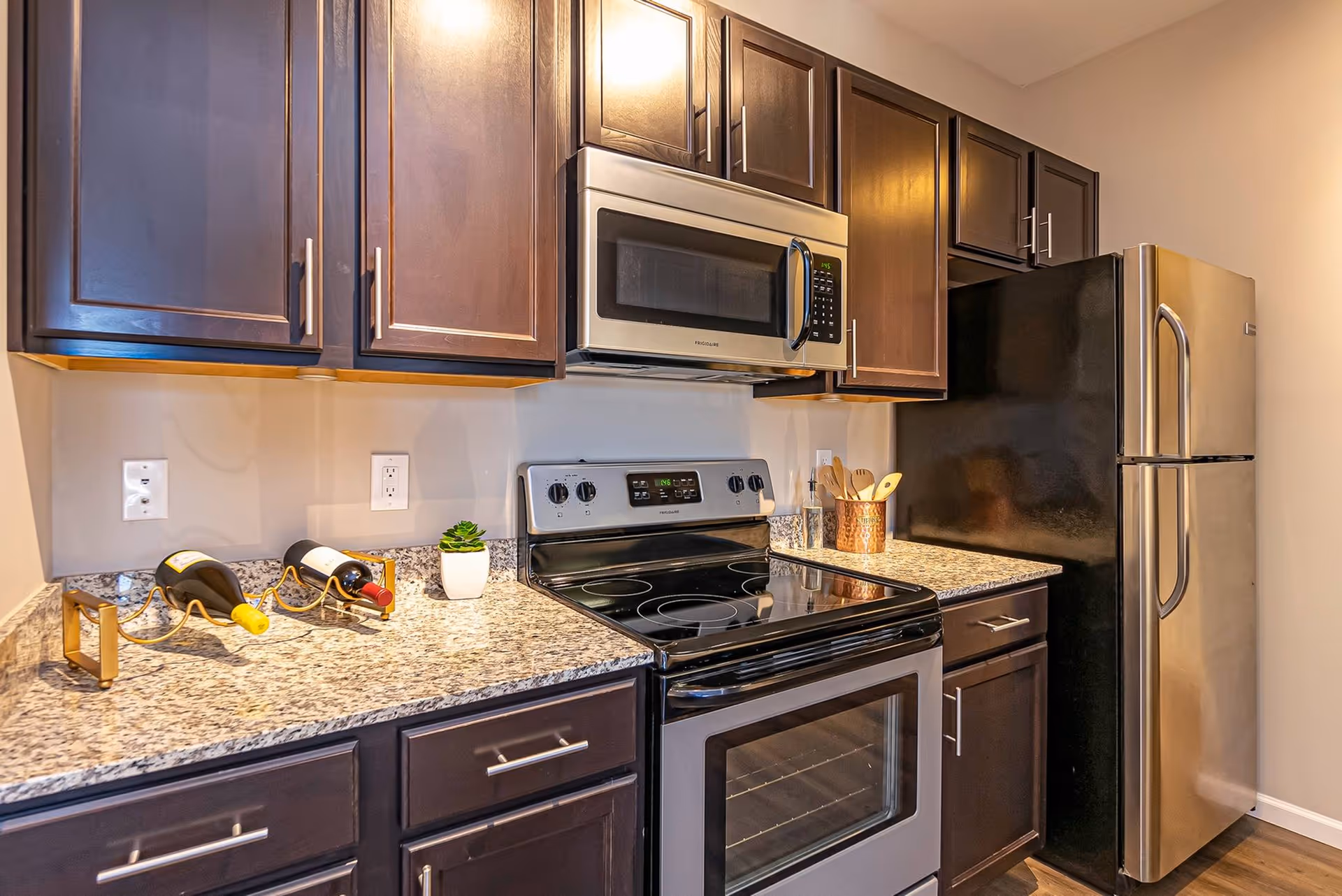 Modern kitchen with dark wood cabinets, granite countertops, a stainless steel microwave above an electric stove, and a stainless steel refrigerator. The countertop has two wine bottles in a gold wine holder, a small potted plant, and a copper container holding wooden utensils.