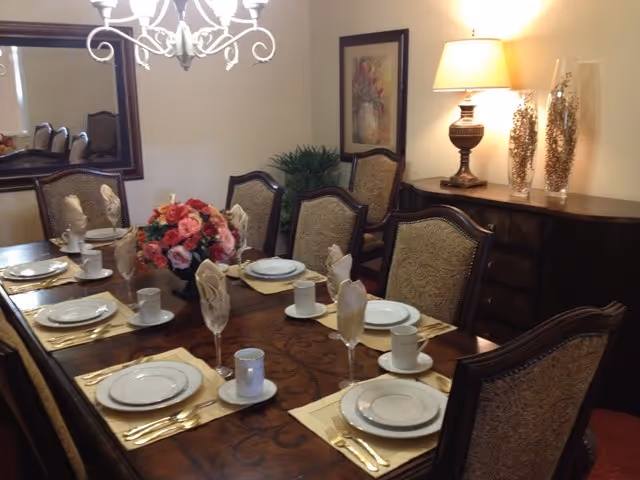 A formal dining room with a wooden dining table set for six people. Each place setting includes a plate, cup, saucer, and folded napkin in a glass. A floral centerpiece decorates the middle of the table. The room features upholstered chairs, a sideboard with a lamp and decorative vases, a framed picture on the wall, and a chandelier overhead.