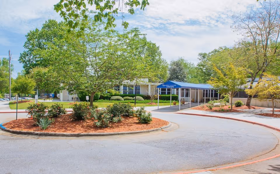 Front exterior of a senior living facility with a circular driveway, landscaped island, trees, and a covered entrance walkway.