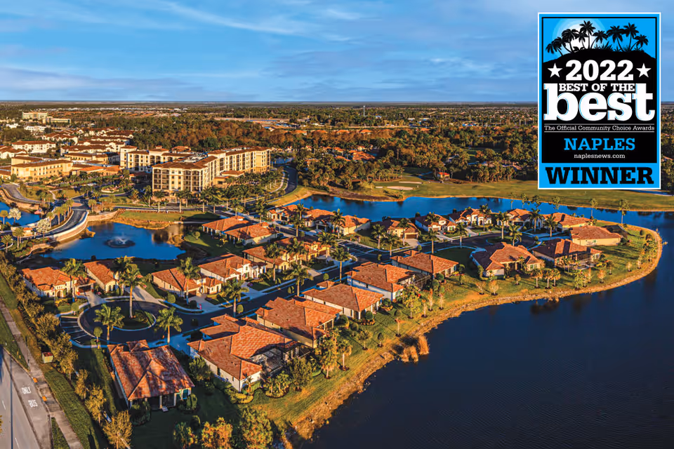Aerial view of a residential community with multiple single-story houses with red-tiled roofs surrounding a lake, with a larger multi-story building in the background and a fountain in a pond. The area is lush with green trees and landscaping under a clear blue sky.