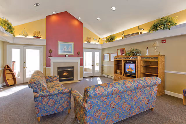 A cozy living room with floral patterned sofas facing a fireplace with a red accent wall. The room has high ceilings with recessed lighting and decorative plants on ledges near the ceiling. There is a wooden entertainment center with a television and various decorative items. French doors with glass panels allow natural light into the room.