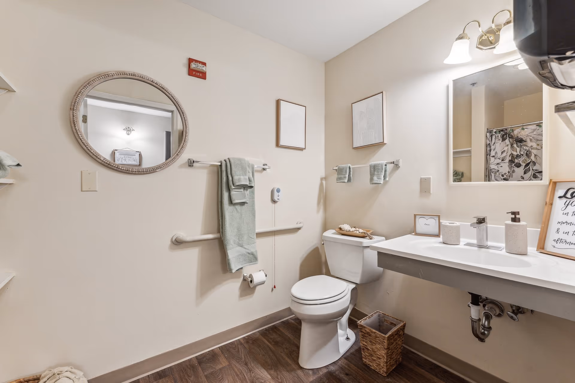Accessible bathroom with a toilet, wall-mounted sink, round mirror, grab bars and neatly hung towels.