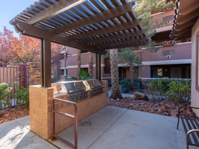 Outdoor barbecue area with two stainless steel grills under a wooden pergola, surrounded by landscaping with rocks, bushes, and trees. The area is part of a senior living facility with multi-story buildings in the background.