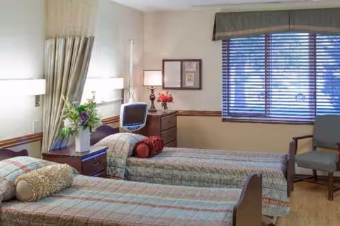 A room with two beds, matching quilts, lamps, and a window with blinds.