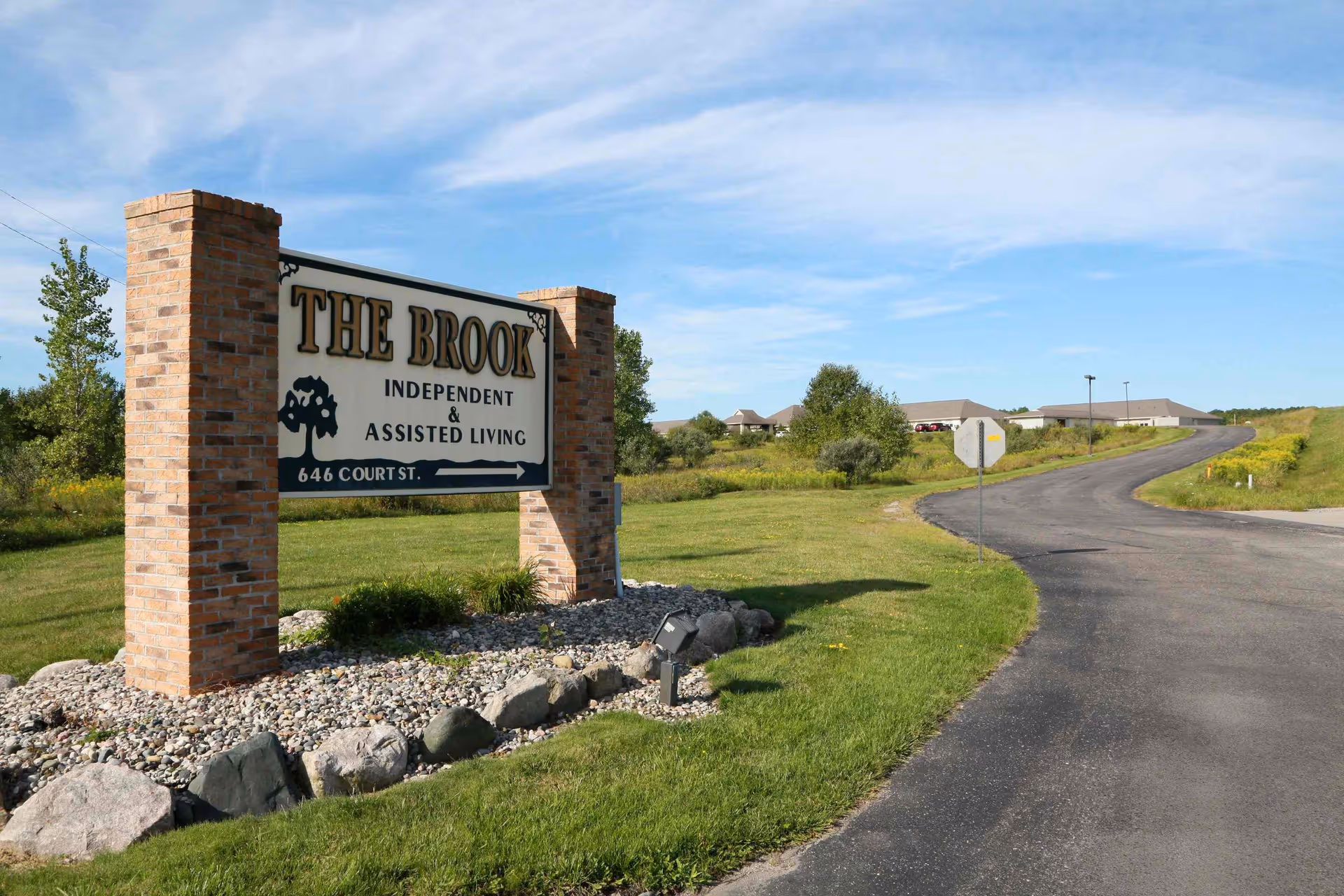 Entrance sign for The Brook independent and assisted living beside a curved driveway leading to the facility buildings.