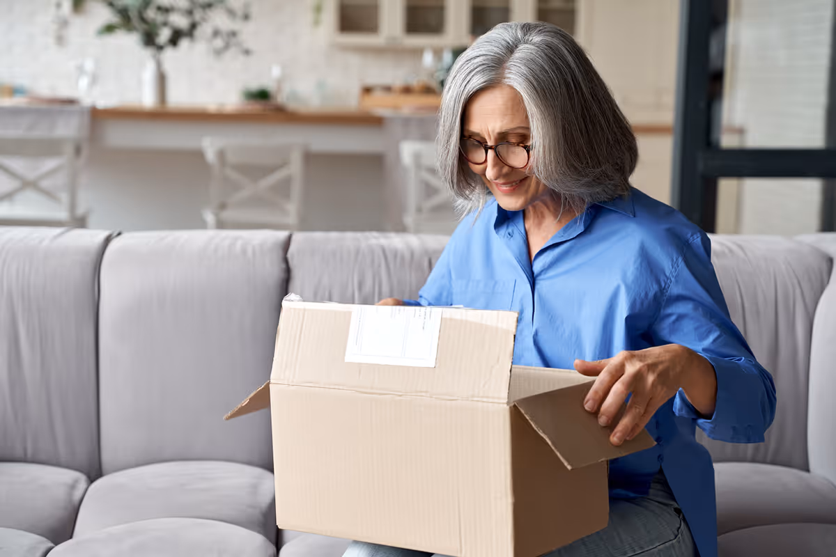 An elderly woman with gray hair and glasses sitting on a light gray couch, smiling as she opens a cardboard box. In the background, there is a kitchen counter with chairs and some plants.