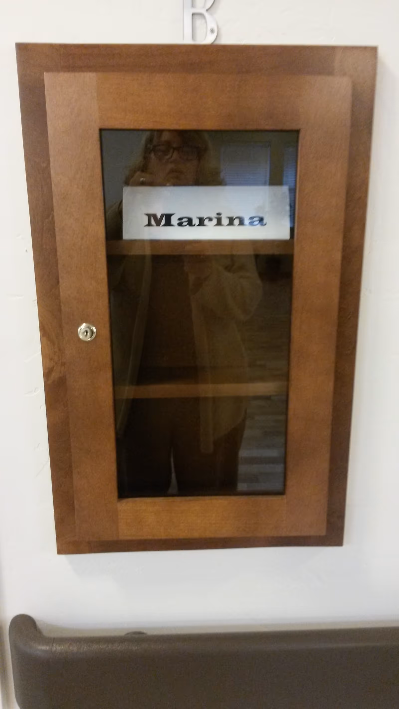 A wooden cabinet with a glass door mounted on a white wall. Inside the cabinet is a white paper with the name 'Marina' printed on it. A person wearing glasses and a light-colored sweater is reflected in the glass of the cabinet.