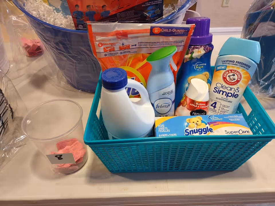 A blue plastic basket containing various household cleaning and laundry products including a bottle of Febreze air freshener, Snuggle fabric softener sheets, Arm & Hammer Clean & Simple deodorant, Renuzit air freshener, and a detergent refill pack. Next to the basket is a clear plastic cup with pink paper slips inside, labeled with the number 8.