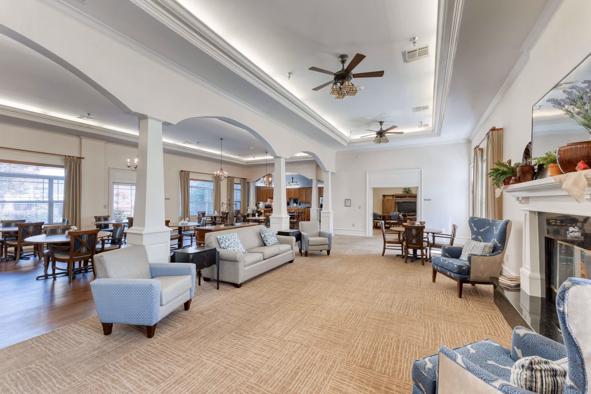 A spacious and well-lit common area in a senior living facility featuring a seating area with armchairs and a sofa, a fireplace with decorative plants on the mantel, and a dining area with multiple tables and chairs near large windows with curtains. The ceiling has recessed lighting and ceiling fans, and the floor is carpeted in the seating area and wood in the dining area.