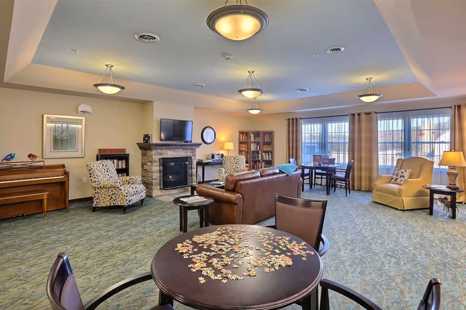 Spacious senior living lounge with seating around a fireplace, a piano, tables with a jigsaw puzzle, and large windows.
