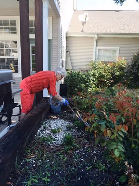 An elderly person wearing a red striped shirt and red pants is gardening in a landscaped area next to a building. They are using gardening tools to tend to plants and bushes near a porch with wooden posts and a barbecue grill.