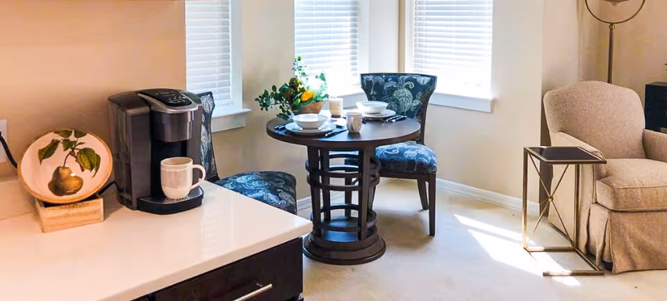 A cozy corner of a senior living facility featuring a small round dining table set for two with blue patterned chairs, a coffee maker on a countertop with a mug, a decorative plate with a pear design, and a beige armchair next to a small side table. Sunlight streams in through two windows with white blinds.