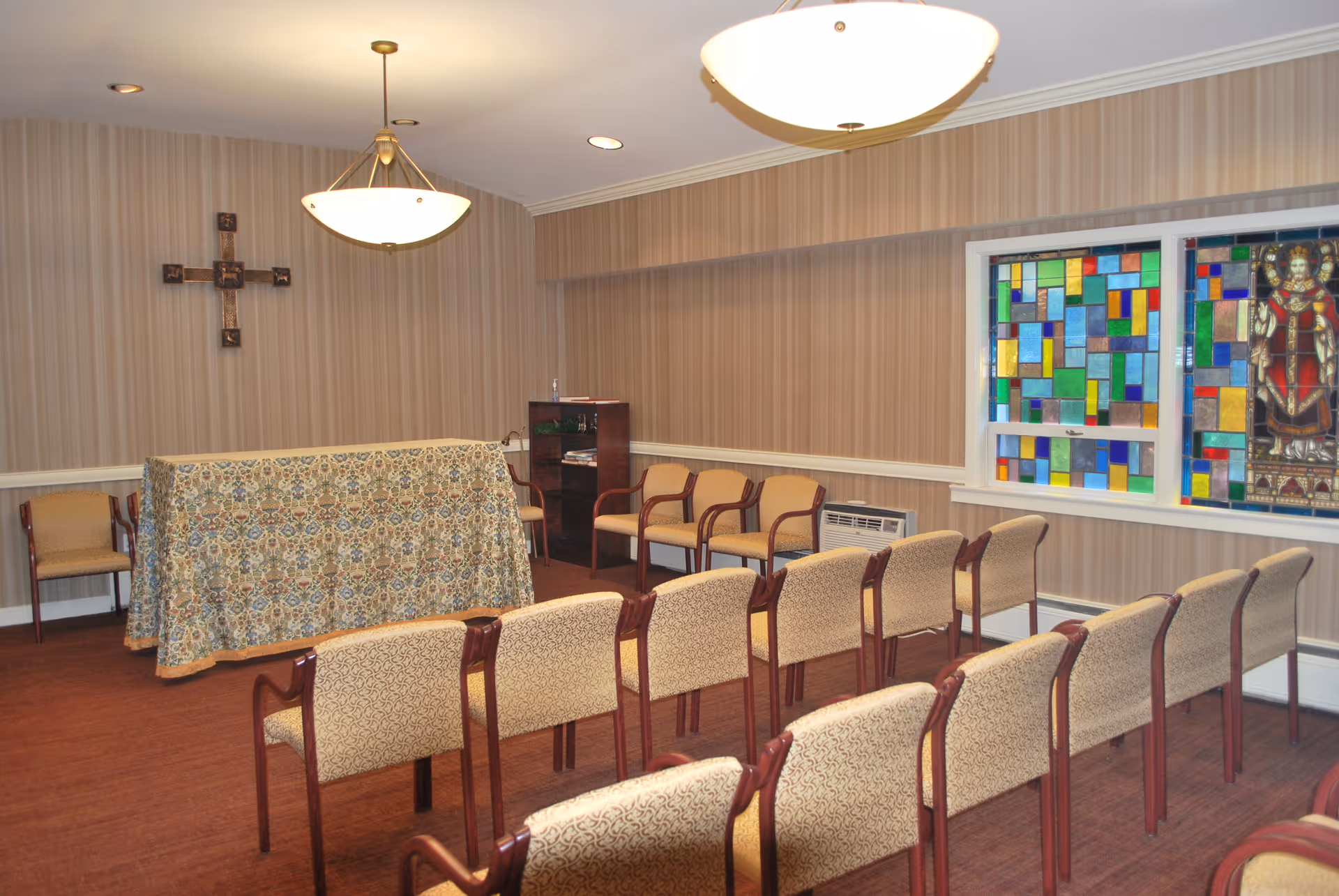 Small chapel-style meeting room with rows of chairs facing an altar covered in patterned cloth and a stained-glass window.