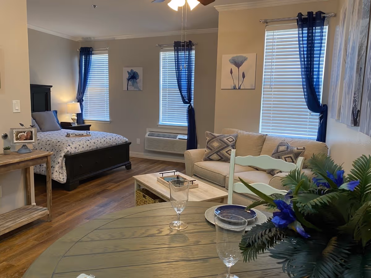 A cozy studio apartment interior featuring a bed with floral bedding and a black headboard, a beige couch with patterned pillows, a wooden coffee table, and a round dining table set with plates and glasses. The room has wooden flooring, three windows with white blinds and blue curtains, and floral artwork on the walls. A small wooden side table with a framed photo and a lamp is next to the bed.