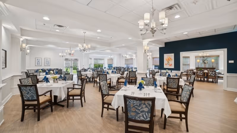 A spacious, well-lit dining room with multiple tables covered in white tablecloths, each set with silverware, glasses, and blue napkins. The chairs have wooden frames with patterned upholstery. The room features chandeliers, large windows letting in natural light, and light-colored wooden flooring. There are framed artworks on the walls and a dark accent wall in the background.