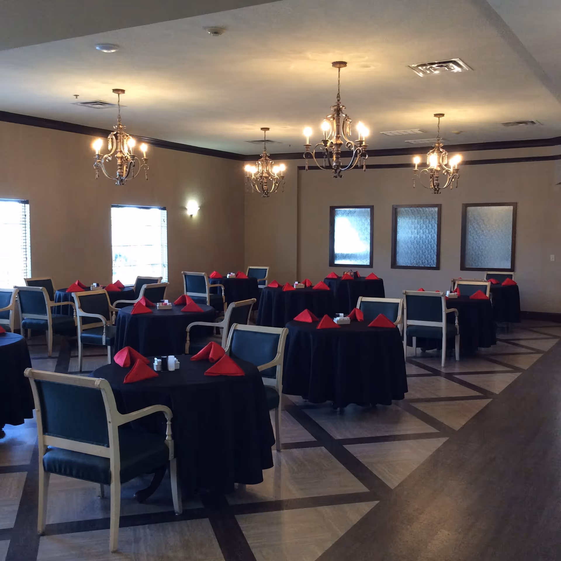 Dining room with multiple round tables covered in black tablecloths, each set with red folded napkins and salt and pepper shakers. The room has wooden chairs with green cushions, three windows with blinds, and four chandeliers hanging from the ceiling providing warm lighting.