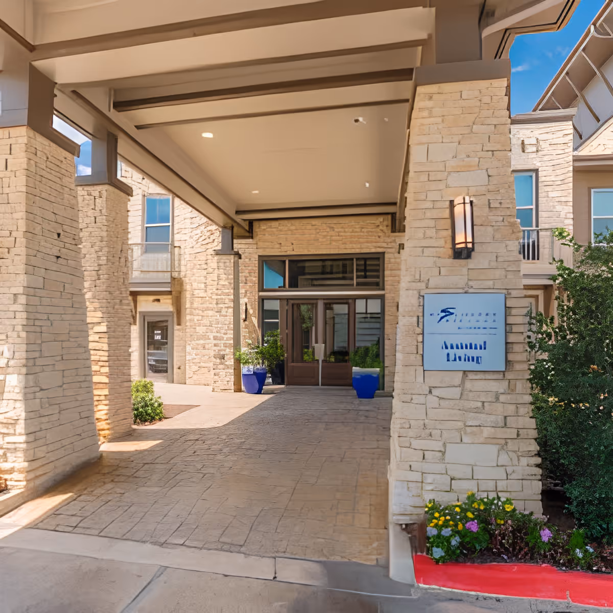 Covered entrance area of a building with stone pillars and walls, glass double doors at the end, potted plants on either side, and a small garden with flowers to the right.