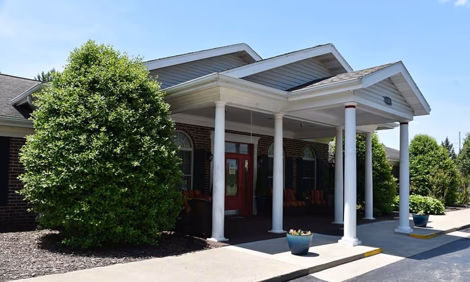 Front exterior view of a single-story brick building with a covered entrance supported by white columns. There are large green bushes on either side of the entrance and a red door in the center. The sky is clear and blue.