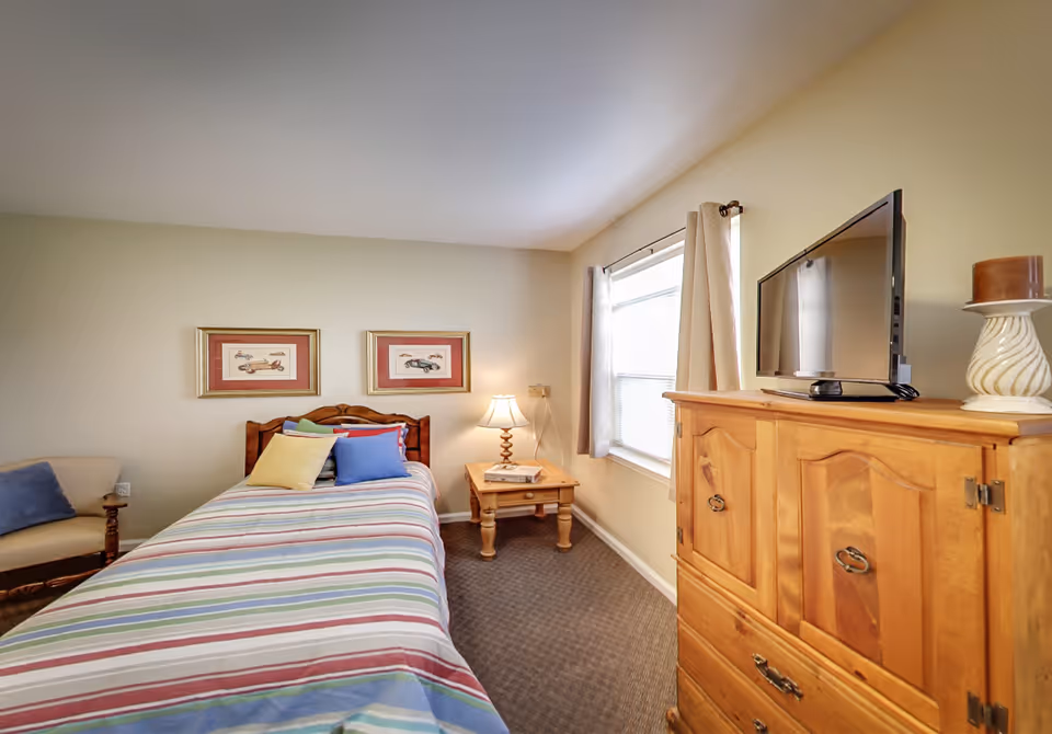A cozy bedroom with a single bed covered in a striped bedspread and several colorful pillows. There is a wooden nightstand with a lamp and a book next to the bed. A wooden dresser with a flat-screen TV and a decorative candle holder is positioned near a window with beige curtains. Two framed pictures hang on the wall above the bed, and a cushioned chair with a blue pillow is partially visible.