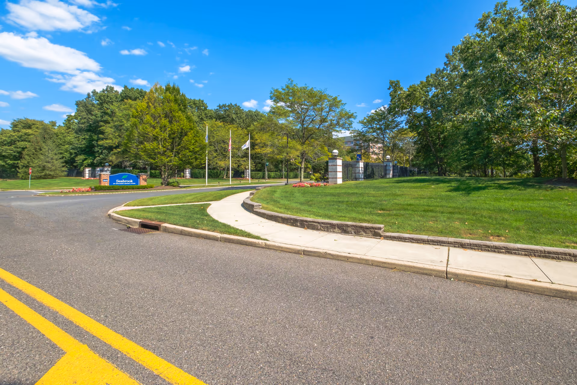 Entrance to Seabrook senior living facility with a curved sidewalk, green lawn, trees, and a blue sign displaying the facility name. Three flagpoles with flags are visible near the entrance under a partly cloudy blue sky.