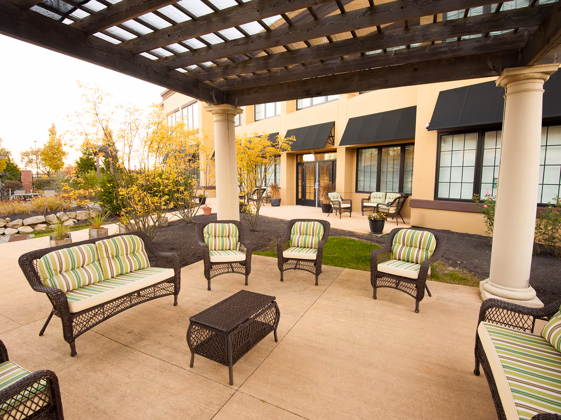 Outdoor patio with wicker sofas and chairs with green-striped cushions under a wooden pergola in front of a building.