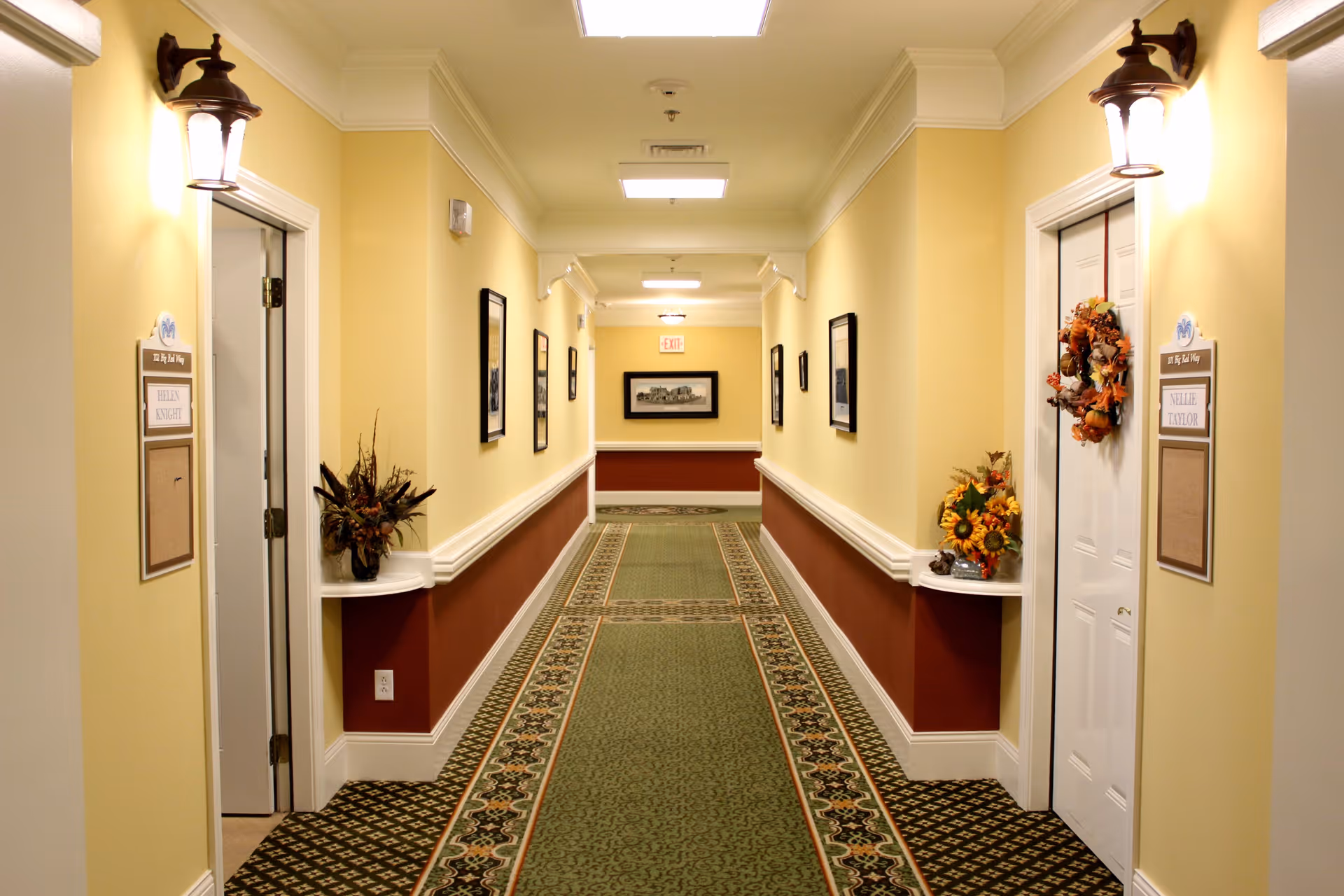 A well-lit interior hallway of a senior living facility with patterned carpet, yellow walls, framed pictures, and doors with nameplates and seasonal wreaths.