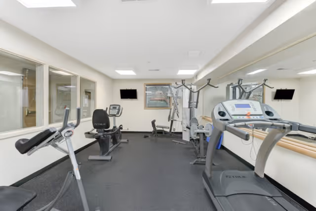 Bright fitness room with treadmills, a recumbent bike, strength machines, mirrors and wall-mounted TVs.