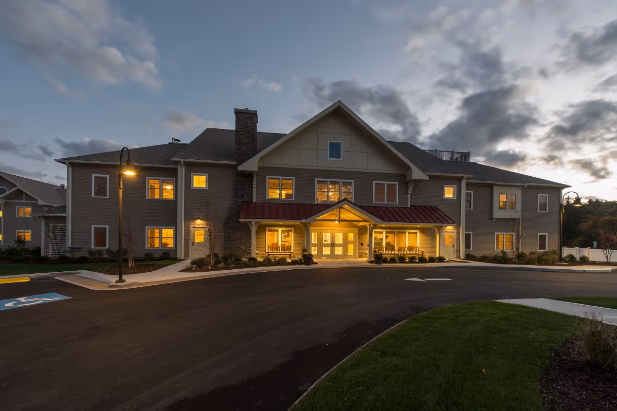 Exterior view of a two-story senior living facility building at dusk with warm lights glowing from the windows. The building has a covered entrance with a red roof, stone chimney, and surrounding landscaping including grass and shrubs. A street lamp is lit near the driveway with a handicap parking space visible.