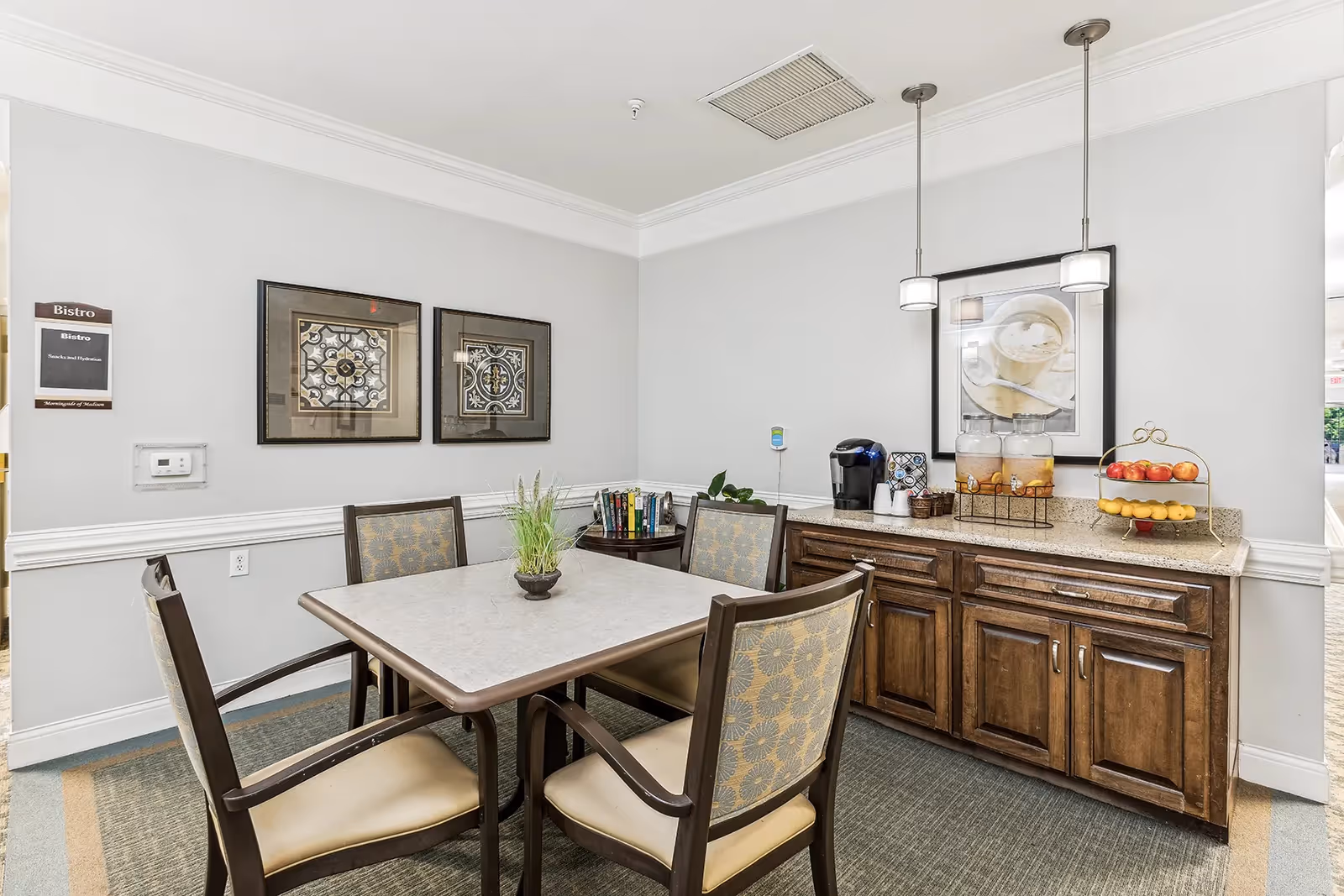 A small dining area with a square table surrounded by four chairs. On the table is a small potted plant. Against the wall is a wooden cabinet with a countertop holding a coffee maker, two beverage dispensers, a basket with cups, and a two-tiered fruit stand with apples and bananas. Above the cabinet hangs a framed picture of a cup of coffee. The walls are light gray with white trim, and two framed decorative artworks are hung on the adjacent wall. A sign on the wall reads 'Bistro'.