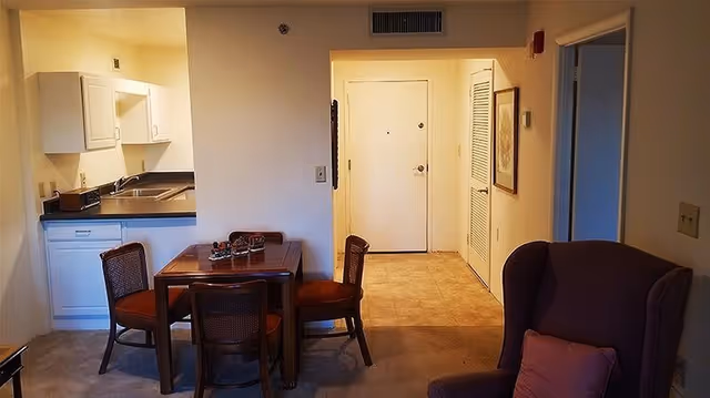 Interior view of a retirement community apartment showing a small dining area with a wooden table and four chairs, a kitchen with white cabinets and a countertop, and an entryway with a closed white door. A purple armchair with a cushion is visible in the foreground on the right side.