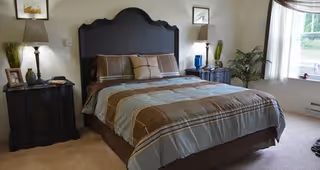 A neatly made bed with a dark wooden headboard in a bedroom. The bed has a brown, blue, and beige striped comforter with matching pillows. On either side of the bed are dark wooden nightstands with lamps, framed pictures, and decorative items. A window with white curtains allows natural light into the room, and there is a green potted plant near the window.