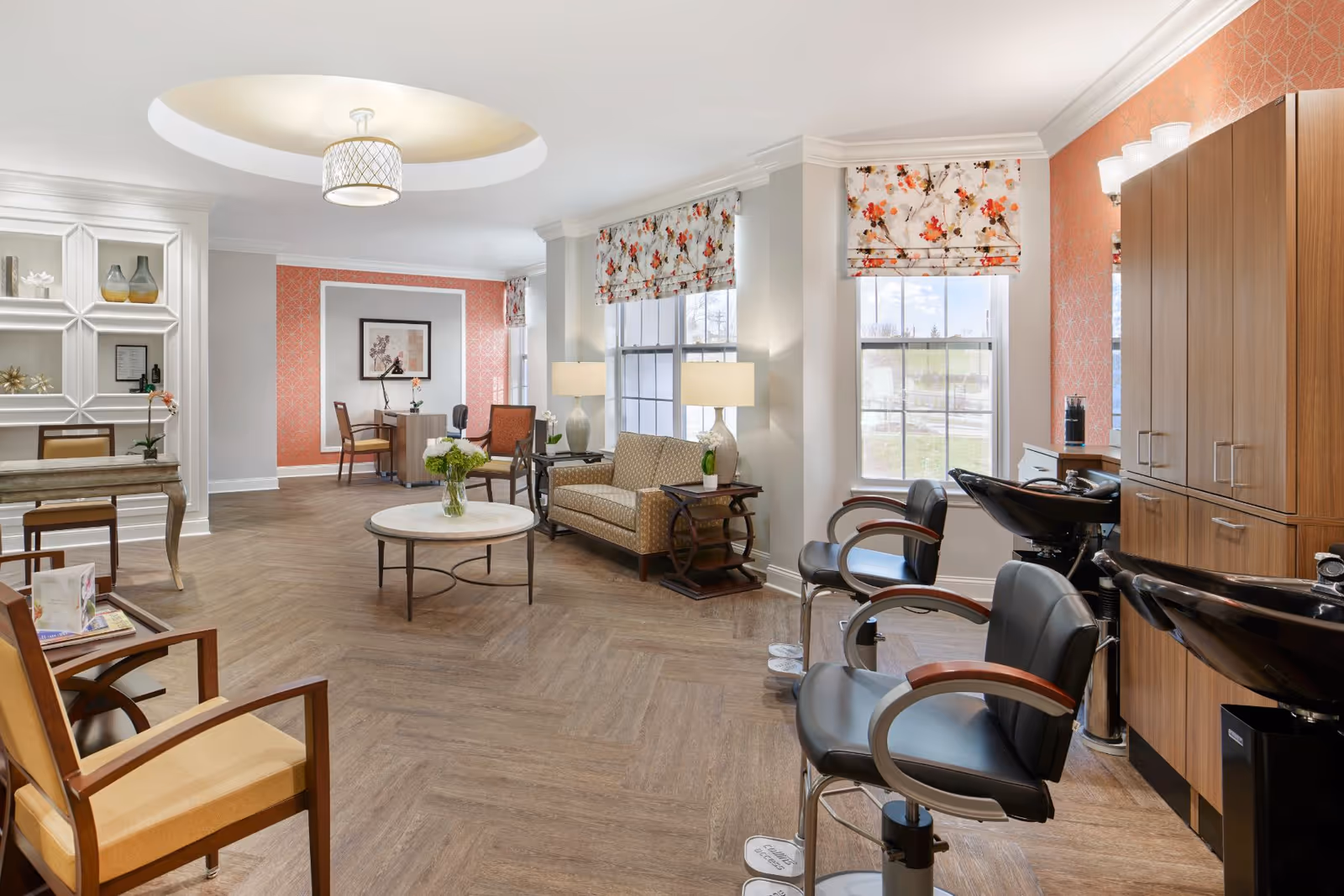 Bright interior common area with seating, a central coffee table, and salon stations with chairs and wash sinks near the windows.