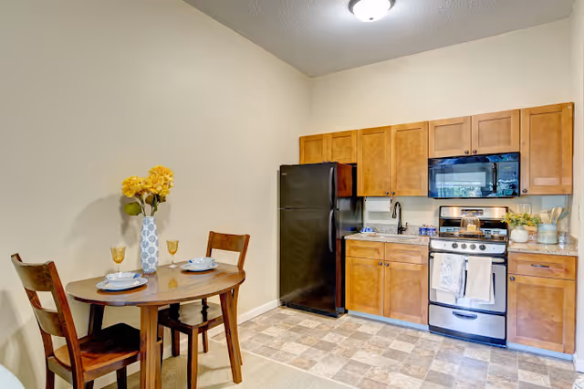 A small kitchen and dining area featuring wooden cabinets, a black refrigerator, a stainless steel stove with a microwave above it, and a round wooden dining table set for two with plates, glasses, and a vase of yellow flowers.