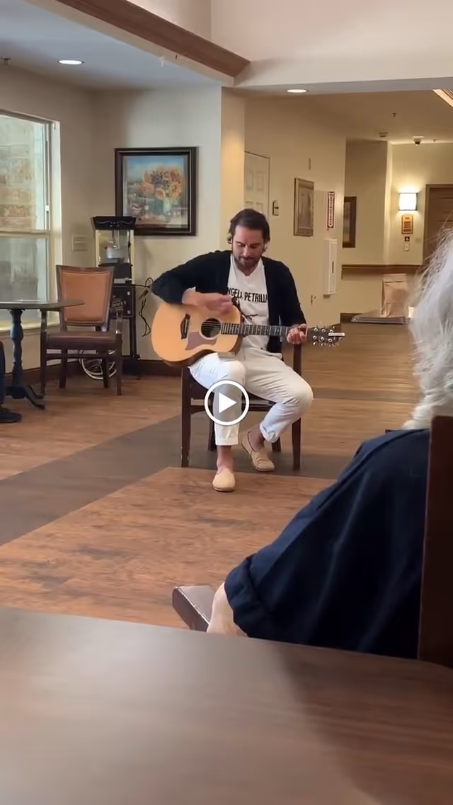 A man is sitting on a chair playing an acoustic guitar in a common area of a senior living facility. The room has wooden flooring, a table with chairs near a window, and framed artwork on the wall. Another person is partially visible in the foreground, watching the performance.