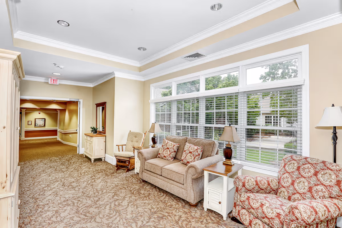 A bright and cozy sitting area in a senior living facility with a beige sofa adorned with patterned cushions, a matching armchair with a red and beige floral pattern, two wooden side tables with lamps, a rocking chair, and large windows with white blinds letting in natural light. The area is carpeted with a patterned design and leads to a hallway with an exit sign above the door.