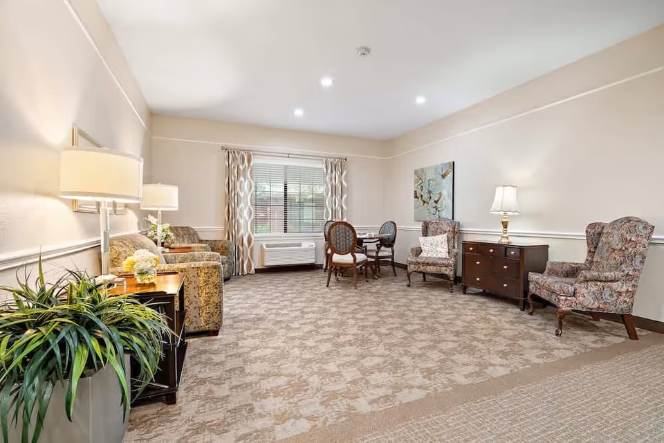 A spacious living room with beige walls and carpeted floor. The room features two floral upholstered armchairs with a small dark wooden dresser and a table lamp between them on the right side. On the left side, there are two patterned armchairs with a side table and a lamp. A small round dining table with four chairs is positioned near a window with patterned curtains. A large green plant is placed in the foreground on the left side.