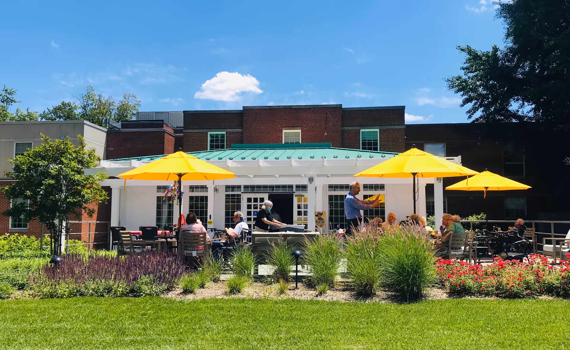 Outdoor patio at a retirement community with people seated under bright yellow umbrellas in front of a brick building.