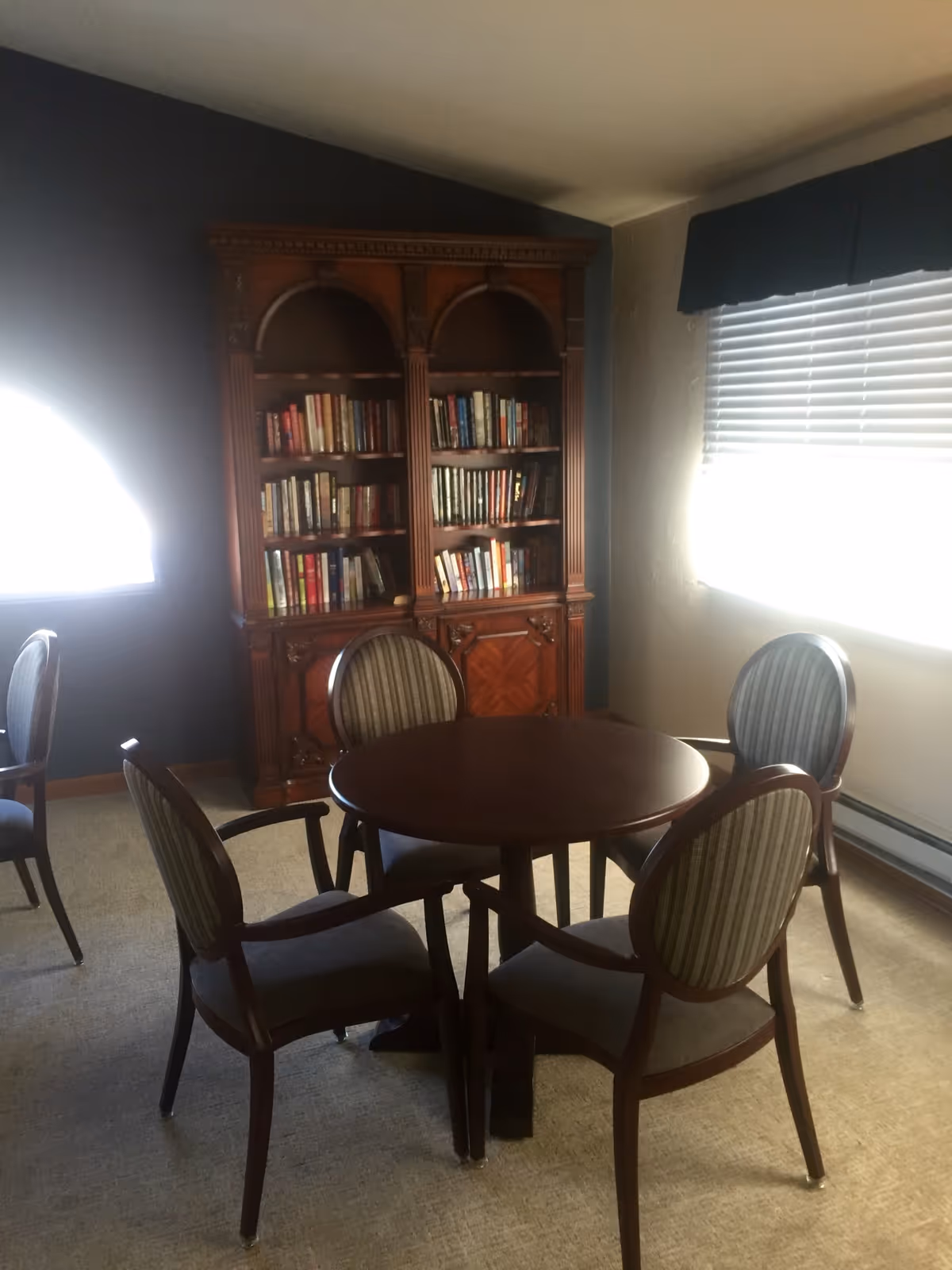 A cozy interior room with a round wooden table surrounded by four upholstered chairs. Behind the table is a large wooden bookshelf filled with books. The room has carpeted flooring and two windows with blinds, allowing natural light to enter.