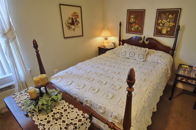 A cozy bedroom with a wooden four-poster bed covered with a white crocheted bedspread and matching pillow. There are two framed floral artworks hanging above the bed. A bedside table with a lamp and a telephone is on one side, and another small table with decorative items is on the other side. A window with sheer white curtains lets in natural light. A wooden bench at the foot of the bed holds candles and a green plant on a crocheted doily.