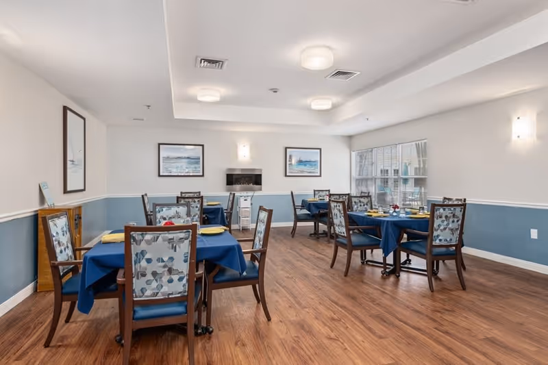 A dining room in a senior living facility with several tables covered with blue tablecloths, each set with yellow napkins and plates. The chairs have patterned upholstery and wooden frames. The room has wood flooring, light blue and white walls, framed artwork, and large windows letting in natural light.