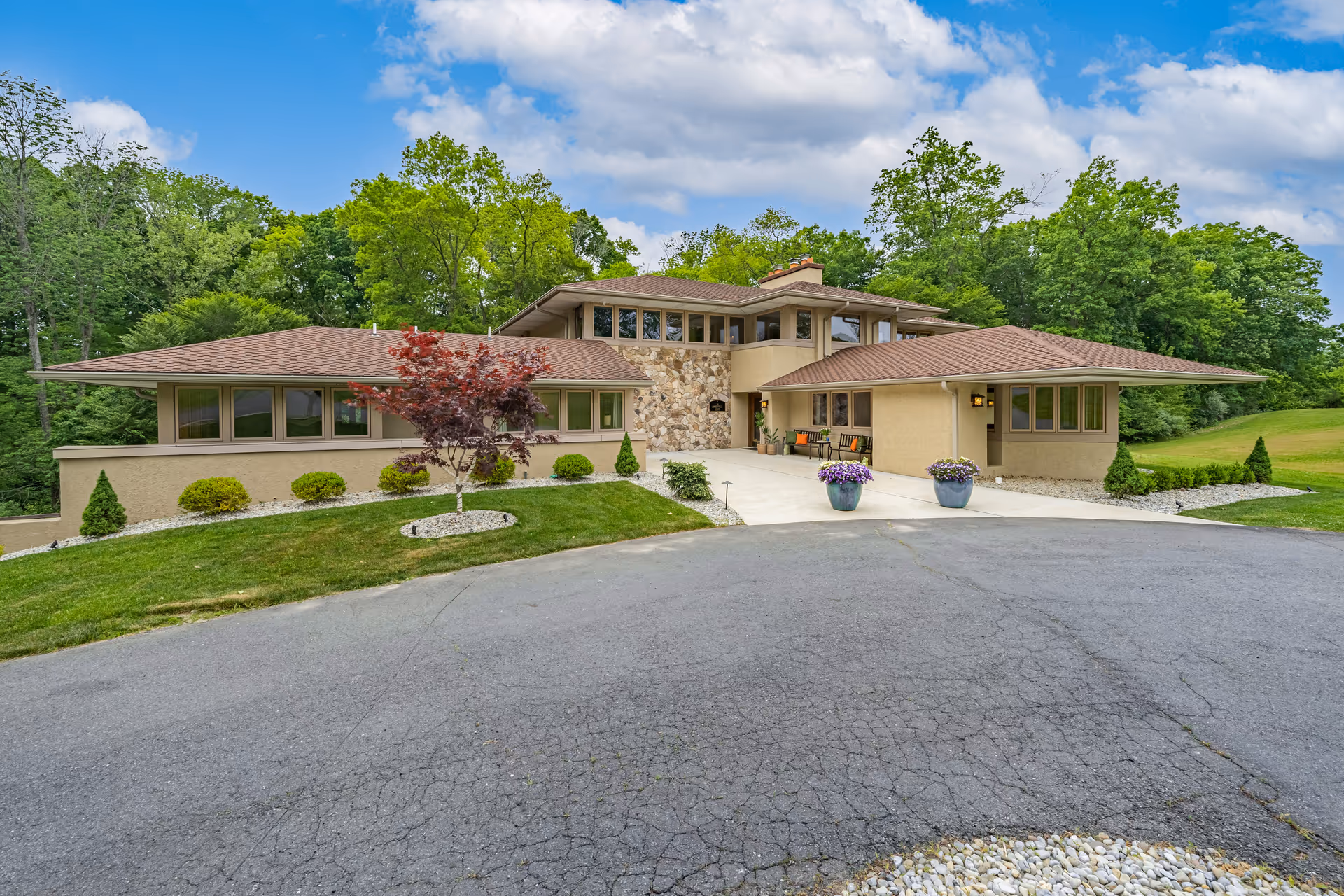 Front exterior view of The Lambertville Estate, a large single-story building with a stone and beige stucco facade, surrounded by green trees and a well-maintained lawn with shrubs and potted flowers under a partly cloudy blue sky.