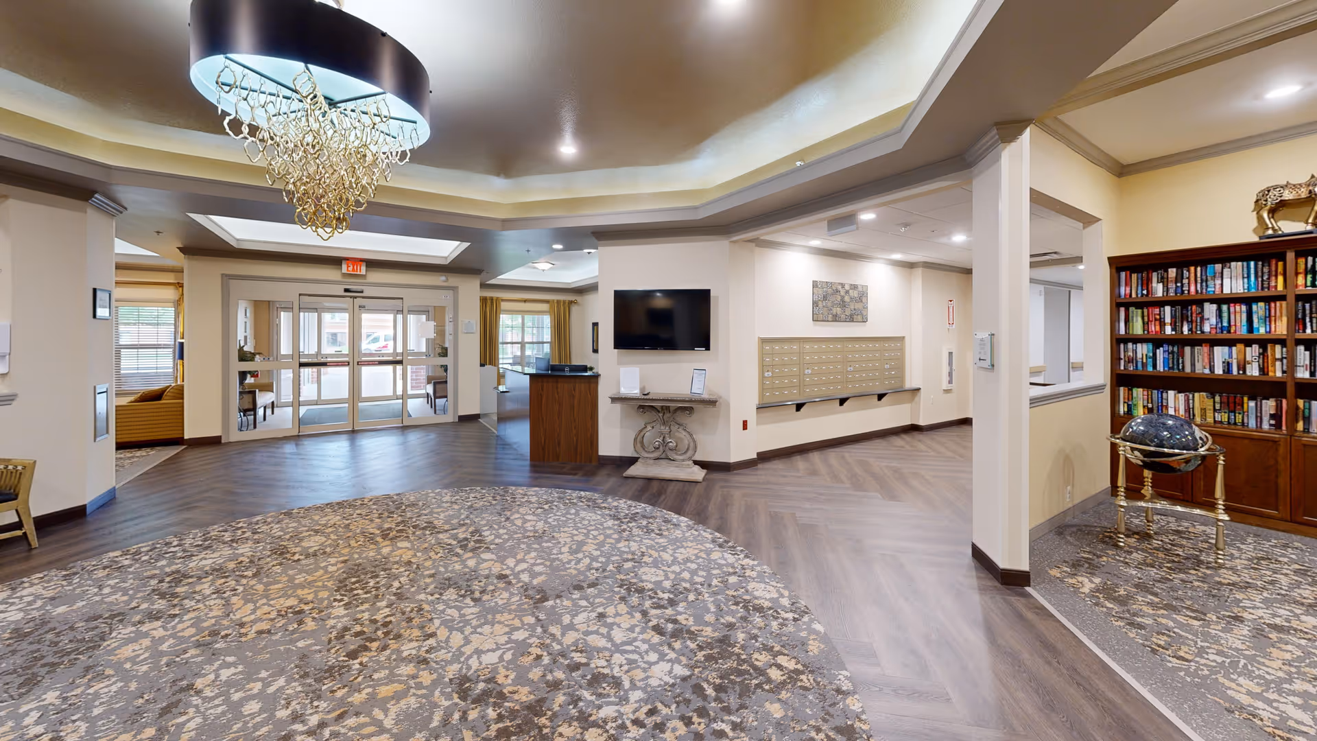 Spacious assisted living lobby with a large patterned rug, chandelier, reception desk, mailboxes, entry doors, and a bookcase.