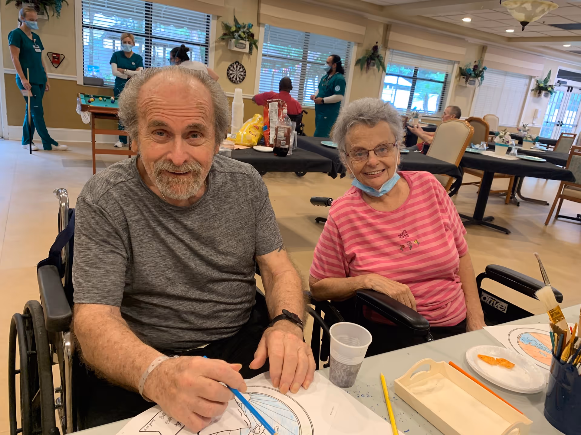 Two elderly individuals sitting in wheelchairs at a table in a nursing facility, engaging in a coloring activity. The man on the left is holding a blue crayon and smiling, while the woman on the right is wearing glasses and a pink striped shirt, also smiling. In the background, several staff members in green uniforms are standing and interacting, with a pool table and dartboard visible in the room.
