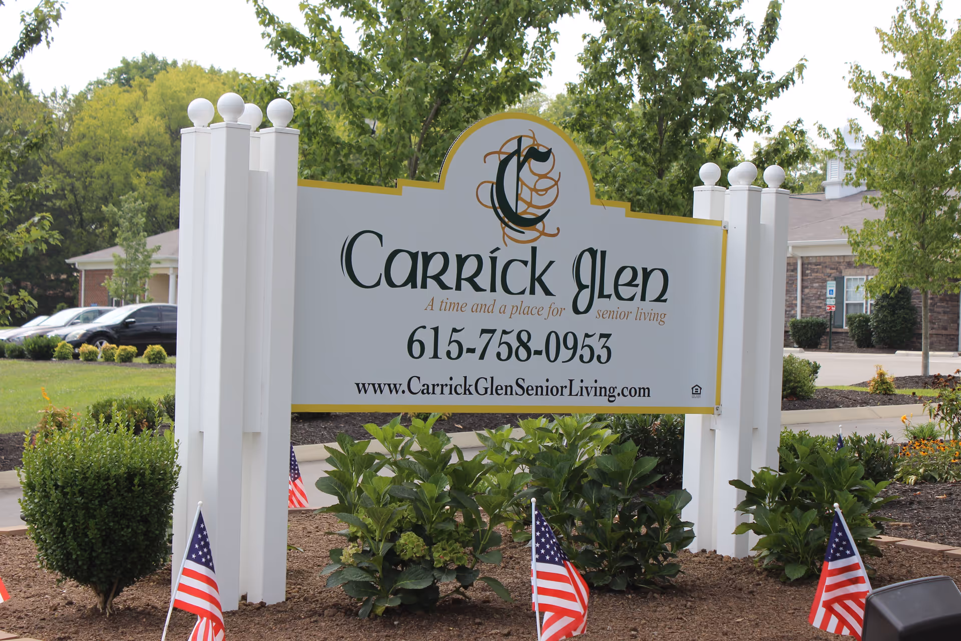 Outdoor sign for Carrick Glen Senior Living with contact phone number and website, surrounded by small bushes and American flags, with trees and a building in the background.